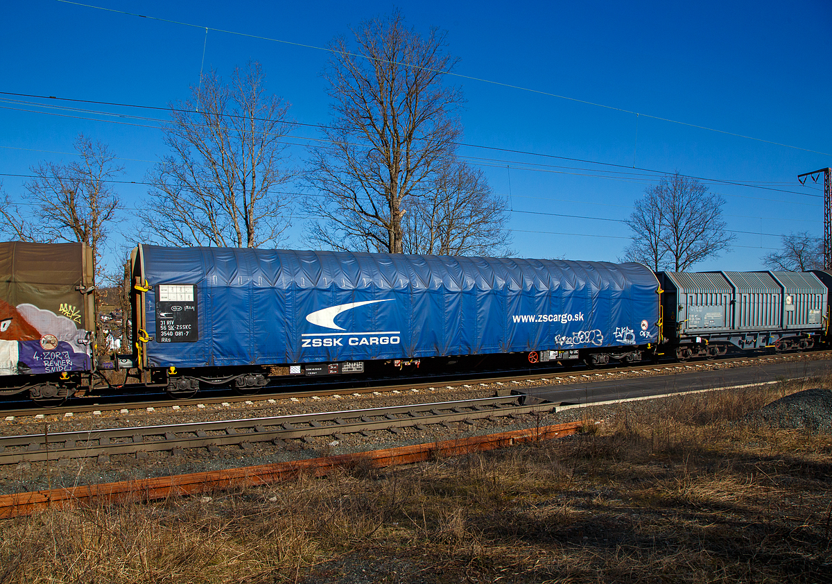 4-achsiger Drehgestell-Flachwagen mit verschiebbarem Planenverdeck, 31 56 3540 081-7 SK-ZSSKC, der Gattung Rils, der slowakischen ZSSK Cargo (Železničná spoločnosť Cargo Slovakia), am 10.03.2022 im Zugverband bei einer Zugdurchfahrt in Rudersdorf (Kr. Siegen).

Die Ausrüstung mit Planen-(Schnell-)Verdeck ermöglicht den Transport von Gütern, für die ein Nässeschutz erforderlich ist. Das Verdeck zeichnet sich durch einfache Handhabung mit Ein-Mann-Bedienung, Leichtgängigkeit und Zentralverriegelung aus. 

Das Planendach wird in geschlossener Stellung manuell verriegelt. Die 4-Punkt-Zentralverriegelung an der Stirnwand ist robust und einfach. Durch die gewählte Anordnung der Betätigungseinrichtung ist das Entriegeln des Planendaches sowohl vom Erdboden als auch von einer Rampe aus möglich. Die beidseitig an jeder Stirnwand angeordneten Verriegelungshaken einschließlich ihrer Betätigungshebel sind über eine drehbar gelagerte Welle miteinander verbunden. Hierdurch wird das Betätigen von nur einer Wagenseite möglich.
Das Planendach lässt sich jeweils zu einem Wagenende hin so weit zusammenschieben, dass jeweils ca. 2/3 des Wagens freigelegt werden kann. 

Die Wagenabdeckung erfolgt mit einer PVC-beschichteten Gewebeplane, die auf 18 Rohrrahmen aufliegt und über Laufwagen in Wagenlängsrichtung verschoben werden kann. Die unteren Laufwagenrollen wirken als Abhebesicherungen. Sie greifen unter die Laufschienenunterkanten und verhindern so ein Abheben der Hauben bei Auflaufstößen oder seitlicher Windbelastung.

TECHNISCHE DATEN:
Spurweite: 1.435 mm (Normalspur)
Anzahl der Achsen: 4 in 2 Drehgestellen
Länge über Puffer: 19.900 mm
Drehzapfenabstand:  14.860 mm
Achsabstand in den Drehgestellen: 1.800 mm
Eigengewicht: 24.360 kg
Ladelänge: 18.500 mm
Ladefläche: 49,3 m²
Maximale Ladegewicht: 55,7 t (Streckenklasse C)
Höchstgeschwindigkeit: 100 km/h (beladen) / 120 km/h (leer)
Kleinster befahrbarer Gleisbogenhalbmesser: R 35 m
Feststellbremse: ja
Bremse: DK-GP (LL) SZ 12
Bremssohle: IB 116
Intern. Verwendungsfähigkeit:  RIV
