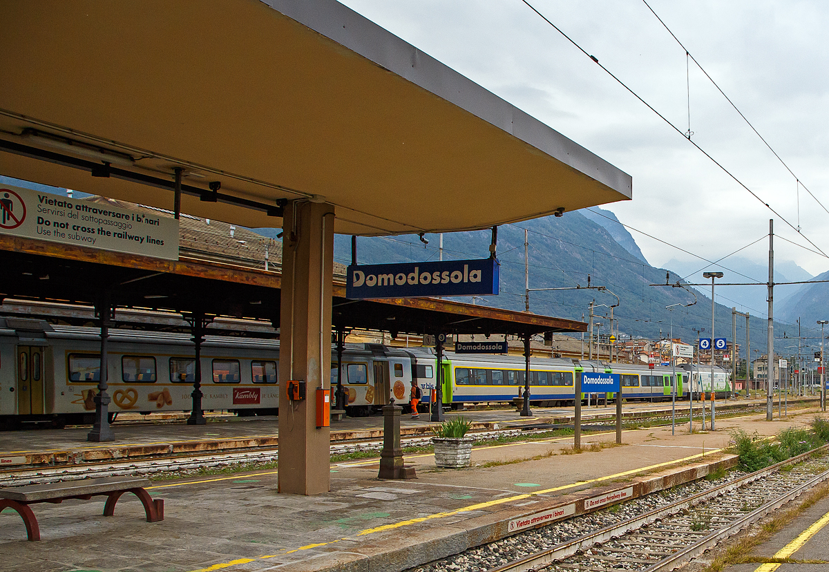 Auf besonderen Wunsch, leider mit vielen Masten über die Bahnsteige hinweg....
Die BLS Werbelok Re 465 008-1  Allegra  (ex Niederhorn) mit Werbung Patenschaft für Berggemeinden steht am 08.09.2021, mit einem EW III Zug u.a. mit dem 1.Klasse Kambly-Wagen. A 50 85 18-34 002-3 CH-BLS, im Bahnhof Domodossola zur Abfahrt nach Brig bereit.
