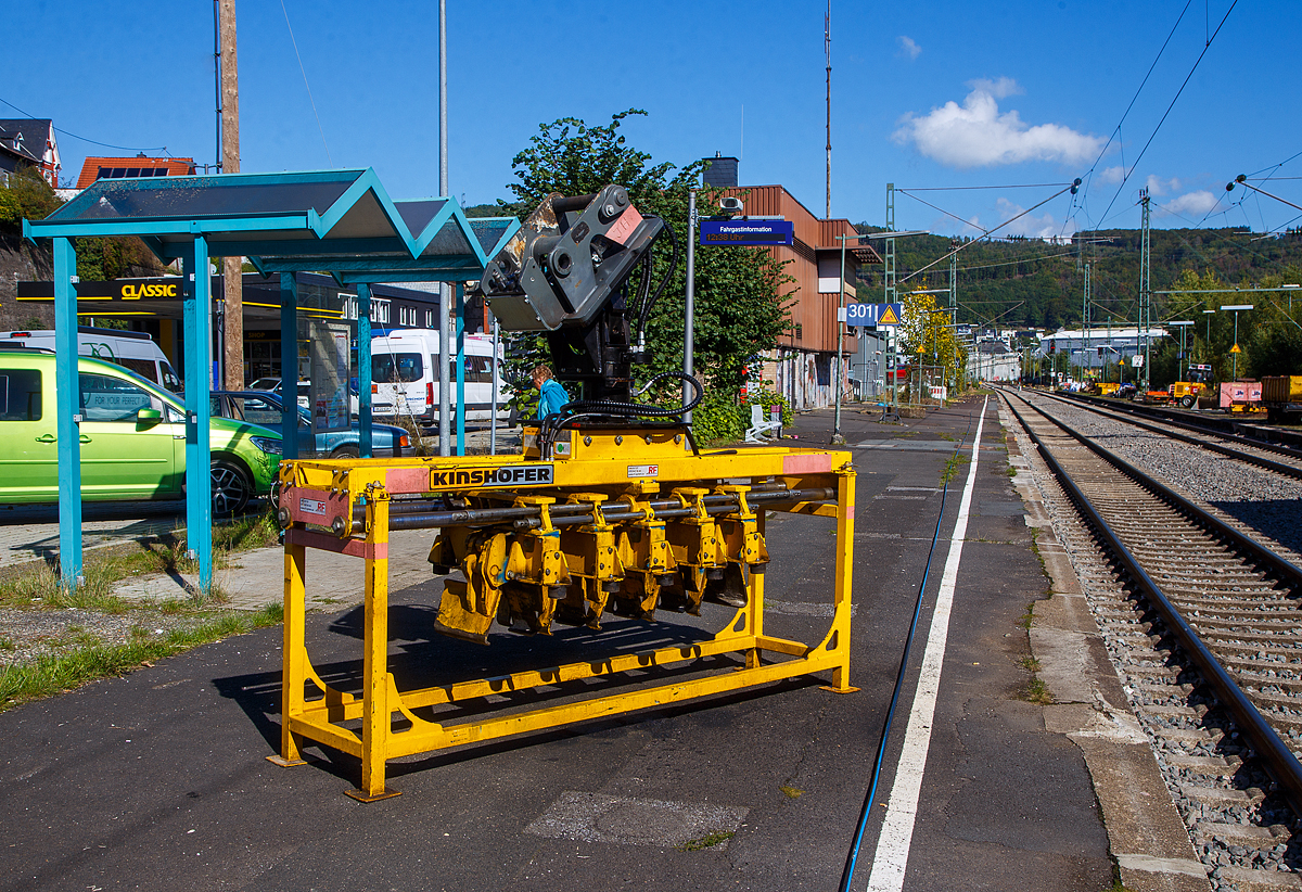 Bahnschwellenleger für Bagger vom Typ RUS05 im Geräteständer, der Joseph Hubert GmbH & Co. KG, am 30.09.2022 abgestellt am Bahnsteig vom Bahnhof Niederschelden (geografisch eigentlich Niederschelderhütte).

Der Hersteller (Baujahr 2020) von dem Bahnschwellenleger ist die KINSHOFER GmbH / RF SYSTEM in (Oberbayern).

Mit dem RUS05 Bahnschwellenleger können Betonschwellen mittig
gegriffen und dann einfach und effektiv positioniert werden, er ist geeignet für den Anbau an Bagger mit 14t bis 24t Dienstgewicht.

Technische Merkmale:
• Greifarme mit Synchronlauf und Untergreifen der Schwellen.
• Effizient: Mehrere Betonschwellen werden gleichzeitig durch hydraulische Greifer mittig geklemmt und anschließend auf das Schotterbett gelegt. Dies spart Zeit im Vergleich zur Einzelpositionierung der Schwellen.
• Sicher: Ein spezielles Halteventil garantiert ein sicheres Halten der Last, selbst bei Druckabfall.
• Variabler Greifbereich: Die Position der Greifarme ist innerhalb des vorgegebenen Bereichs verstellbar.
• Flexibilität: Für fast alle taillierten Bahnschwellentypen. Höhenanschlag je Schwelle einstellbar.
• Für enge Bereiche, da die Schwellen mittig gegriffen werden.
• Präzise Positionierung dank aufgebautem 10t-Drehmotor mit 4 Öldurchführungen und Schlauchschutz.
• Inklusive Geräteständer.
• Einstellbarer Schwellenabstand: 600mm / 620mm / 650mm

Der RUS05 Bahnschwellenleger ist ein hoch-spezialisiertes Gerät zum Greifer und Positionieren von Bahnschwellen nach einer bestimmten Anordnung. Der Abstand der Schwellen kann abhängig von den Anforderungen schnell und einfach angepasst werden. Die Bahnschwellen werden mittig von den voreingestellten, hydraulischen Greifarmen geklemmt und auf dem Schotterbett positioniert. Im Vergleich zur individuellen Positionierung der Bahnschwellen wird viel Zeit eingespart. So gut wie alle Arten von taillierten Bahnschwellen können sicher geklemmt werden, dank integriertem Lasthalteventil auch bei plötzlichem Druckabfall.

Der RUS05 wird inklusive praktischem Geräteständer ausgeliefert, zur einfachen Lagerung und Transport des Gerätes wenn nicht am Trägergerät montiert. Als Variante gibt es auch den RUS2 05 Bahnschwellenleger, dieser ist wie der RUS05 speziell für das Verlegen von fast allen Schwellentypen konzipiert, allerdings werden Bahnschwellen bei dieser Variante über die Enden geklemmt und nicht mittig. Beide Schwellenleger sind mit einem 10t-Drehmotor ausgestattet und können 5 Schwellen gleichzeitig greifen.

TECHNISCHE DATEN Bahnschwellenleger RUS05:
Greifbereich: 150 bis 265 mm
Gewicht: 1.300 kg
Länge: 2.820 mm
Höhe: 1.553 mm
Breite: 825 mm
Anzahl der Greifzangen: 5
Backenbreite: 330 mm (Schwellenbreite 300 / 320 mm)
Max. Gewicht  Einzelschwelle 350 kg (5 x 350 = 1.750 kg Gesamtlast)