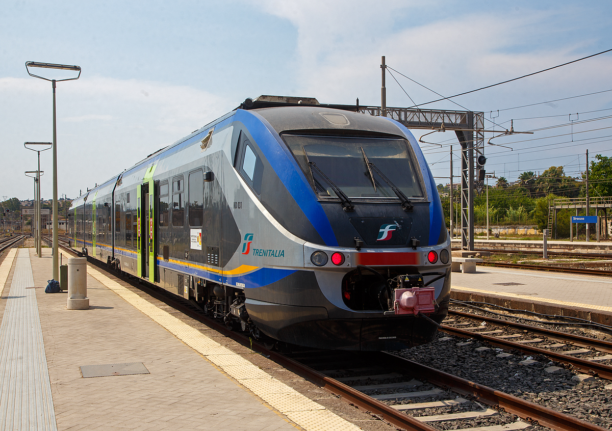 Der dreiteilige Minuetto Diesel MD 031 der Trenitalia steht am 19.07.2022 im Bahnhof Syrakus (Stazione di Siracusa), als Regionalzug R 12938 nach Vittoria, zur Abfahrt bereit. Er f�hrt via Fontane Bianche, Avola, Noto, Pozzallo, Sampieri, Ragusa, Donnafugata und Comiso, so f�hrt er auch an den s�dlichen Zipfel von Sizilien.

Diese dreiteiligen Diesetriebz�ge der italienischen Baureihe ALn 501 / Ln 220 / ALn 502 (MD - Minuetto Diesel) sind die Diesel Variante der Alstom-Coradia-Meridian-Familie, �hnlich wie die ME - Minuetto Elettrico (ALe 501 / Le 220 / ALe 502). Die italienischen Minuetto MD und ME sind die Mutter der Produktfamilie der Alstom Coradia Meridian, von diesen wurden sp�ter auch Triebz�ge f�r Deutschland und Nordeuropa abgeleitet. 

Von dieser Diesel Variante ALn501 / Ln220 / ALn 502 (MD) wurden 114 St�ck, zwischen 2003 und 2009, von Alstom Ferroviaria S.p.A. (ex Fiat Ferroviaria) in Italien gebaut. Von der elektrischen Variante ALe 501 / Le 220 / ALe 502 (ME) wurden 136 St�ck zwischen 2003 und 2010 gebaut. Beide Varianten sind als dreiteilige Triebz�ge ausgef�hrt. Der Minuetto ist komfortabler klimatisierter Triebwagenzug der auf dem Alstom Coradia Meridian basiert. 

Er wurde von Giorgetto Giugiaro entworfen und wurde speziell f�r den italienischen Markt entwickelt. Gebaut wurden sie in Italien von Alstom Ferroviaria in den italienischen Produktionsstandorten. Die Endmontage und Gesamtprojektmanagement geschah im ehemaligen Fiat Ferroviaria Werk in Savigliano. Die Drehgestelle kommen aus dem Alstom Werk Salzgitter in Deutschland.

Wie auch die anderen Z�ge der Coradia-Plattform, sind die Wageneinheiten fest miteinander verbunden und k�nnen nur in au�ergew�hnlichen Wartungs- oder Pannenf�llen getrennt werden. Ein Triebzug ist durchg�ngig niederflurig und barrierefrei.

Die Antriebsk�pfe ruhen jeweils auf einem Triebdrehgestell (konventionelle Enddrehgestelle) und st�tzen sich zum anderen auf einer H�lfte eines Jakobs-Drehgestelles ab. Der Mittelwagen ruht beidseitig jeweils auf einer H�lfte eines Jakobs-Drehgestelles. Die Triebz�ge sind Scharfenberg-Kupplungen ausgestattet, mit der bis zu drei Z�ge in einem Zug verbunden werden k�nnen. Ein Dieseltriebzug kostet rund 3,45 Millionen Euro pro Fahrzeug, 150.000 Euro mehr als die Elektro-Version.

Die beinahe 52 m langen Fahrzeuge bieten 23 Sitzpl�tze in der 1. Klasse und 98 Sitzpl�tze in der 2. Klasse an. Der Antrieb erfolgt auf die beiden Enddrehgestelle, w�hrend die beiden Jakobs-Drehgestelle ohne Antrieb sind. Jedes Triebdrehgestell wird von im Unterflur eingebauten gedrosselten IVECO-V8-Dieselmotor mit Common-Rail-Direkteinspritzung, vom Typ IVECO V8 FVQE 28 angetrieben. Die Kraft�bertragung vom Motor aufs Drehgestell geschieht hydraulisch, durch ein hydromechanisches Getriebe mit hydrodynamischer Bremse vom Typ Voith T 212 bre.

TECHNISCHE DATEN der MD - Minuetto Diesel:
Spurweite:  1.435 mm (Normalspur)
Achsformel:  Bo' 2' 2' Bo'
L�nge �ber Puffer:  51.900 mm
Drehzapfenabstand: 14.650 mm / 13.650 mm /14.650 mm
Achsabstand im Trieb-Drehgestell:  2.400 mm
Achsabstand im Lauf-Drehgestell:  2.700 mm
Treibraddurchmesser:  850 mm
Breite. 2.950 mm
H�he:  3.820 mm
Eigengewicht:  100 t
H�chstgeschwindigkeit:  130 km/h
Motoren: 2 St�ck V8-Dieselmotor mit Common-Rail-Direkteinspritzung, Typ Iveco V8 FVQE 28
Getriebe: Voith T 212 bre (hydromechanisch mit hydrodynamischer Bremse)
Leistungs�bertragung: hydraulisch
Nennleistung (gedrosselt): 2 x 560 kW (760 PS) bei 2.100 U/min
Leistung ungedrosselt: 2 x 720 kW (980 PS)
Sitzpl�tze:  23 in der 1. Klasse und 98 in der 2 Klasse
Kupplung: Scharfenberg