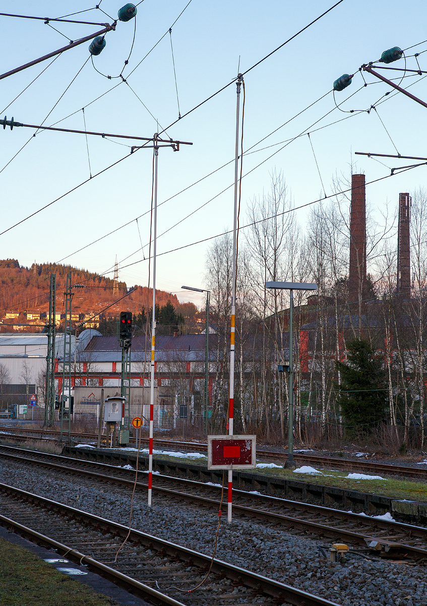 
Niederschelderhütte (zu Mudersbach, DB mäßig nur Niederschelden) den 13.02.2018:Die Oberleitung ist geerdet und stromlos....

Die Erdungsstangen sind ein gehangen, am Haken oberen Ende der Stange ist das Kabel angeschlossen, welches mittels Schienenklemme an das Gleis angeschlossen ist und somit ist die Oberleitung geerdet. An der vorderen Stange ist zudem eine Sh 2 - (Schutz-)Halttafel befestigt. Diese Tafel gebietet Halt und ersetzt ein haltzeigendes Hauptsignal. Nachts leuchtet an der Tafel zusätzlich ein rotes Licht.

Eine Erdungsstange, auch Erdungsstab, ist eine isolierende Stange mit deren Hilfe die Oberleitung von Hand geerdet werden kann. Am oberen Ende ist ein Haken fürs Erden fest angebracht. Das daran befindliche Kupferkabel wird dann an die Schiene (Gleis) an geklemmt, um so den Erdschluss herstellen. Mit der Erdungsstange wird im Rahmen von Installations- oder Wartungsarbeiten an freigeschalteten Oberleitungen ein Kurzschluss der Fahrleitung hergestellt, um Stromunfälle durch elektrische Spannungen zu verhindern.

Die Erdungsstange ist eine rote/weiße (oder gelbe) Stange aus glasfaserverstärkten Kunststoff (GFK). Je nach Einsatzzweck kann die Stange als Teleskopstange ausgeführt sein oder aus mehreren Einzelteilen zusammensteckbar sein. Sie ist so leichter zu transportieren und vor der Witterung geschützt in Taschen unterzubringen. Durch das Zusammenstecken aus einzelnen Elementen werden Längen von mehreren Metern erreicht. Die Erdungsstange ist dabei auf eine solche Länge zusammenzubauen, dass das zu erdende Anlagenteil erreicht wird und Sicherheitsabstände nicht unterschritten werden.

Die korrekte Vorgehensweise beim Erden mit Erdungsstangen ist eine angepasste Abfolge wie bei den Fünf Sicherheitsregeln:
1. Freischalten der Fahrleitung. (Oberleitung ausgeschaltet)
2. Gegen Wiedereinschalten sichern
3. Spannungsfreiheit feststellen: (Teilweise haben Erdungsstangen auch Spannungsprüfer eingebaut)
4. Erdungskabel Schienenfuß befestigen
5. Erden und kurzschließen

So wird sichergestellt, dass Personen nicht durch eventuell auftretende Kriechspannungen oder gar durch das unbeabsichtigte Einschalten der Fahrleitung gefährdet werden.
