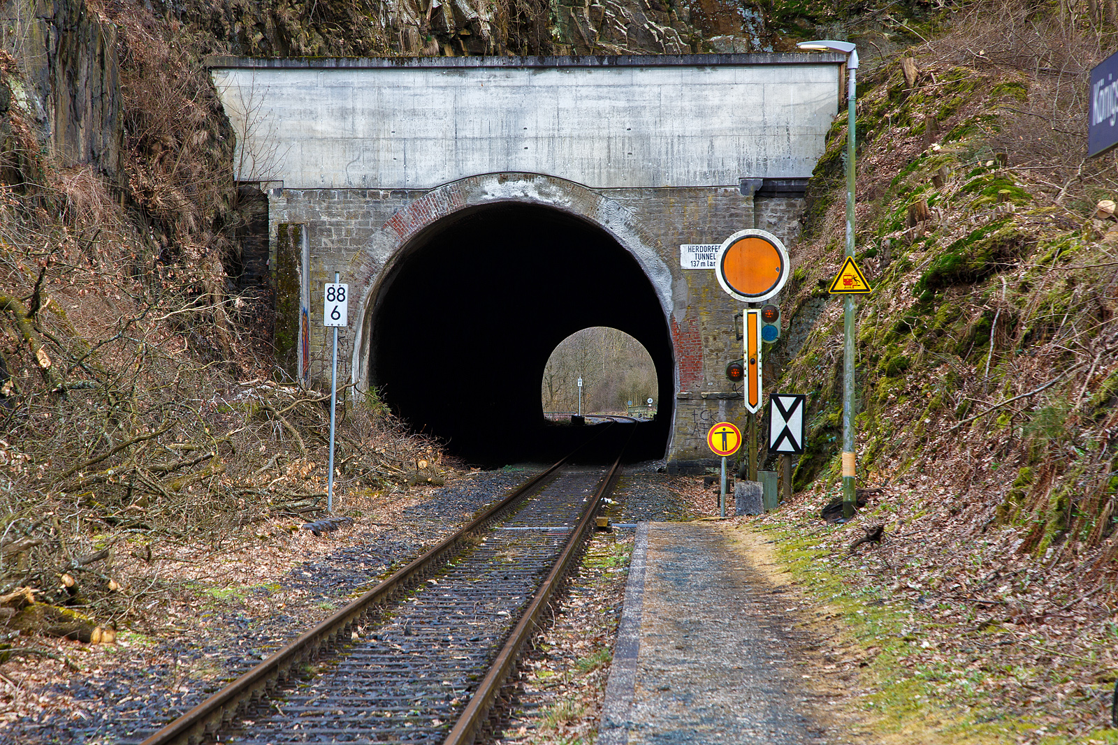 Der 137 m lange Herdorfer Tunnel hier am 18.03.2023 vom Hp K�nigsstollen gesehen. 

Davor das Vorsignal (f�rs Einfahrtsignal Bf Herdorf) Vr 0 - Halt erwarten! Die gelbe Scheibe mit wei�em Rand und gelbe Pfeil zeigt nach unten. Nachts leuchten auch die beiden gelben Lampen. Vor dem Signal die Ne 2 – Vorsignaltafel. Die Vorsignaltafel zeigt dem Lokf�hrer, da� das Signal, vor dem sie steht, Vorsignalfunktion hat, d.h., den Begriff eines nachfolgenden Hauptsignales vorank�ndigt. 
