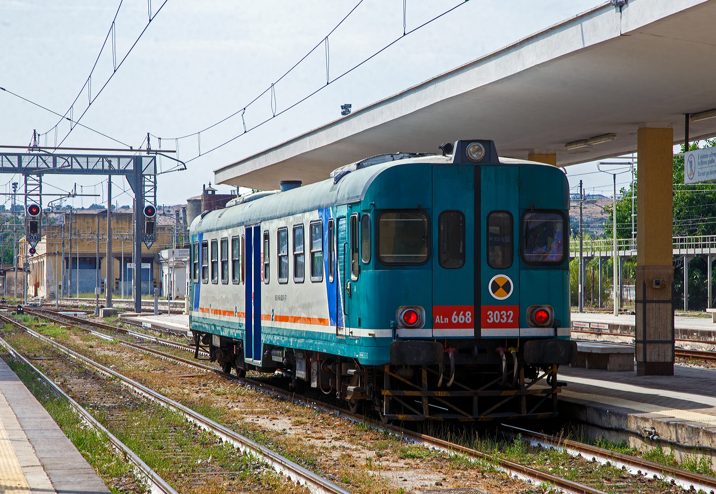 Der Dieseltriebwagen ALn 668 3032 (95 83 4668 332-8 I-Ti) der Trenitalia (100-prozentige Tochtergesellschaft der FS) fährt am 19 Juli 2022 in Bahnhof Syrakus (Stazione di Siracusa) ein und wird als Regionalzug als R 12946 nach Augusta bereitgestellt. Die roten Lichter täuschen hier.  

Der Dieseltriebwagen (Verbrennungstriebwagen) wurde 1980 von der Officine Meccaniche Calabresi O.ME.CA S.p.A. in Reggio Calabria gebaut und an die FS - Ferrovie dello Stato Italiane (Italienische Staatseisenbahnen) geliefert. Mit der Aufteilung in einzelne Betriebsgesellschaften der FS im Jahr 2000 kam der VT zur heutigen Tochtergesellschaft Trenitalia.

Die O.ME.CA. (deutsch Kalabrische Mechanische Werkstätten) war ein Hersteller von Schienenfahrzeugen mit Sitz in der Stadt Reggio Calabria, später gehörte sie zur AnsaldoBreda S.p.A., im Laufe der Zeit führte der Auftragsrückgang zu einer Verkleinerung der gesamten AnsaldoBreda-Gruppe und seit Februar 2015 gehört sie zur Hitachi Rail Italy S.p.A.. Die Stadt Reggio Calabria liegt an der Südspitze der italienischen Apenninhalbinsel (Stiefelspitze) und somit an der Ostseite der Straße von Messina gegenüber von Messina (Sizilien).

Geschichte der ALn 668:
Zu Beginn der 1950er Jahre war der Dieseltriebwagenbestand der italienischen Staatsbahnen für jene Dienste, für den er eigentlich ausgelegt war, praktisch unbrauchbar geworden. Zudem verursachte die große Zahl der verschiedenen Typen einige Schwierigkeiten beim Unterhalt und bei der Beschaffung von Ersatzteilen.

Zur Modernisierung des Reiseverkehrs auf dem sizilianischen Schmalspurnetz der FS wurden ab Januar 1950 Triebwagen des Typs RALn 60 eingesetzt. Bei der Konstruktion dieser Bauart konnten technische Erfahrungen gesammelt werden, die in der Folge auch für die regelspurigen (normalspurigen) Dieseltriebwagen von FIAT verwendet werden konnten.

Im Jahre 1954 wurde dann das Projekt eines standardisierten Dieseltriebwagens aufgenommen, welcher – gestützt auf die Erfahrungen mit den beiden bereits im Betrieb befindlichen Triebwagenserien – die Probleme im Dieselfahrzeugpark der FS lösen sollte. Der neue Triebwagen wurde auf 68 Sitzplätze ausgelegt, davon 8 erster und 60 zweiter Klasse. Gemäß dem Klassifikationssystem der FS wurde er als Leichter Dieseltriebwagen „Automotrice Leggera a Nafta 668“ (Leichter Dieseltriebwagen 668), kurz ALn 668 bezeichnet. Die Verdoppelung der Anfangsziffer „66“ wurde eingeführt, um Baureihen mit Vielfachsteuerung kenntlich zu machen.

Dank der breitgefächerten Verwendungsmöglichkeiten dieser Fahrzeuge und der mehr als 700 Exemplare, welche die italienischen Staatsbahnen in 30 Jahren anschafften, wurde diese Bauart vor allem ab den 1960er Jahren zum eigentlichen Rückgrat des italienischen Regionalverkehrs und erst in neuerer Zeit, durch die Minuetto, fast ganz auf Nebenbahnlinien verdrängt.

Projektierung und Anforderungen an das Fahrzeug:
Im Jahre 1954 bestellten die FS bei der Fiat Ferroviaria die ersten drei Prototypen der Serie: Sie erhielten die Nummern 1401 bis 1403.

Nach den gemischten Erfahrungen mit den großen einmotorigen Triebwagen ALn 990 und Aln 880 orientierte man sich beim Entwurf der neuen Fahrzeuge an die Erfolge mit den Vorkriegs-Triebwagen, den „Littorine“ (ALn 40, ALn 56, ALn 80 usw.), welche mit zwei Motoren ausgestattet waren. Es wurde auch der seit der Vorkriegszeit eingetretene technische Fortschritt mitberücksichtigt und die Technik gegenüber den Littorine wesentlich verbessert.

Die Entscheidung, die neuen Triebwagen mit zwei Motoren auszustatten, bot verschiedene Vorteile: Die beiden unabhängigen Traktionsgruppen mit weitverbreiteten Dieselmotoren erlaubten es, die Kosten für den Antrieb relativ tief zu halten, das Vorhandensein von zwei Treibachsen verbesserte die Adhäsion und die Ersatzteile waren günstiger und einfacher zu bevorraten. Alle mechanischen Teile sind einfach, leicht und wartungsarm ausgeführt. Zudem waren die Dieselmotoren aus der FIAT LKW-Massenproduktion was weiter die Kosten reduzierte.

Gegenüber den Vorkriegstriebwagen neu entwickelt wurde die Steuerung einschließlich der Kupplungsbetätigung, welche nunmehr elektrisch erfolgt. Damit konnte die von Straßenfahrzeugen bekannte Pedalbedienung aufgegeben werden. Ebenfalls eine Neuerung stellte die Verwendung von Lkw-Motoren für Triebwagen dar. Die wichtigste Neuerung betraf jedoch den Einbauort der Motoren. diese wurden nicht mehr im Kasten eingebaut, sondern befinden sich als „Unterflurmotor“ nunmehr unterhalb des Kastens. Damit konnten die Probleme der Instabilität bei hoher Geschwindigkeit und der Übertragung von Vibrationen des Zuges während der Fahrt auf heikle mechanische Teile, welche bei den Vorgängermodellen Quelle von Beschädigungen und häufigen Reparaturen war, gelöst werden.

Die Unterfluranordnung des Fahrmotors war auch das Merkmal der damals neuen Aln 668 Triebwagen, um maximalen Platz für Fahrgäste und Servicepersonal zu schaffen. Diese Technik setzte weltweit Maßstäbe im italienischen Eisenbahnbau, so dass der Einsatz des Horizontalzylindermotors Anfang der 1950er Jahre von den maßgeblichen internationalen Eisenbahnzeitschriften oft als „italienische Lösung“ bezeichnet wurde, die heute Standard ist (heute meist als Power-Packs).

Die Wagen erhielten Seitenpuffer und Regel-Schraubenkupplungen.

Von den Diesel-Triebwagen vom Typ FS ALn 668 wurden von 1951 bis 1981 insgesamt 787 Stück in 12 Serien bzw. drei Generationen gebaut. Mit der dritten Generation, der Serie 1200 und 3000 kehrten die FS wieder zu Mitteleinstiegen zurück, behielten jedoch die größere Wagenlänge der Serie 1900 bei. Durch einen vergrößerten Einstiegsbereich konnte die Ein- und Ausstiegszeit insgesamt verkürzt werden. Die Stirnseiten wurden verändert, das Dach ist an den Wagenenden nicht mehr heruntergezogen. Von dieser Dieseltriebwagen Serie vom Typ FS ALn 668.3000 (ALn 668 Serie 3000) wurden zwischen1980-1981 von FIAT Ferroviaria Savigliano und Officine Meccaniche Calabresi (O.ME.CA), insgesamt 40 Stück, gebaut.

FS Aln 668 – Serie 3000 – der 3.Generation:
Die FS ALn 668.3000-Triebwagen, auch bekannt als ALn 668 Baureihe 3000, gehören zur dritten Generation der ALn 668-Gruppe, die in chronologischer Reihenfolge aus den Baureihen 1900, 1000, 1200, 3000, 3100 und 3300 besteht.

Die Baureihe/Serie 3000 (FIAT Typ 7241 S) ist die aufgeladene Version der Baureihe 1200 (FIAT Typ 7241), die sich durch ihre Höchstgeschwindigkeit von 130 km/h, anstatt der 110 km/h, unterscheidet.

Bau der Serie 3000:
1978 wurden 120 Triebwagen des Typs ALn 668.3000 der FS bestellt. Die Serie blieb jedoch aufgrund der Entscheidung, die Mehrfachtraktionsmöglichkeit auf drei statt zwei Triebwagen zu erweitern, auf 40 Einheiten begrenzt. Diese Entscheidung führte 1980 zur Schaffung der neuen Serie 3100 mit den verbleibenden 80 Triebwagen.

Der Bau der vierzig Triebwagen des Typs ALn 668.3000 wurde dann zu gleichen Teilen zwischen FIAT Ferroviaria und Officine Meccaniche Calabresi (O.ME.CA), aufgeteilt. So wurden die ALn 668.3001-3020 von FIAT Ferroviaria in Savigliano (im Piemont) 1980 und die ALn 668.3021-3040 (wie dieser hier) von der O.ME.CA S.p.A. in Reggio Calabria 1980 und 1981 gebaut und geliefert.

Merkmale:
Die Triebwagen ALn 668.3000 verfügen über eine Kapazität von 68 Sitzplätzen, 8 Sitzplätze in der 1.Klasse und 60 (heute 57) in der 2.Klasse. Die Triebwagen und wurden von zwei Dieselmotoren mit einer Nennleistung von jeweils 147 kW angetrieben, die später auf 170 kW erhöht wurde. Diese ermöglichen eine Höchstgeschwindigkeit von 130 km/h und eine Reichweite von ca. 600 km. 

Die Triebwagen ALn 668.3000 können zusammen mit den Steuerwagen Ln 664.1400 gefahrenwerden und sind für den Mehrfachtraktionsbetrieb in Doppeltraktion geeignet.


Unterschiede zur 1200er-Serie: 
Der ALn 668.3000 unterscheidet sich von der 1200er-Serie nur durch den aufgeladenen IVECO 8217.32-Dieselmotor und die daraus resultierende Dimensionierung der Getriebekomponenten in Abhängigkeit von der höheren entwickelten Leistung, sowie höher Höchstgeschwindigkeit von 130 km/h.

Motor und Getriebe:
Der Motor der 3000er-Serie nutzt die aufgeladenen IVECO 8217.32-Motoren, die ab der 1900er-Serie verbaut wurden. Ihre gesteigerte Leistung ermöglicht eine Höchstgeschwindigkeit von 130 km/h ohne Achs-Getriebewechsel und mit der gleichen Achsübersetzung wie bei den 1900er- und 1000er-Serien.

Der aufgeladene Motor erforderte außerdem den Einsatz eines größeren Hydraulikgetriebes, einer Zweischeibenkupplung und die Verlegung der Schalldämpfer vom Dach in spezielle vertikale Auspufföffnungen an den Seiten des hinteren Vorraums, wie bei den Vorgängermodellen der 1900er- und 1000er-Serie.

TECHNISCHE DATEN der ALn 668 Serie 3000 (3. Generation):
Baujahre: 1980/81
Gebaute Anzahl: 40
Spurweite: 1.435 mm (Normalspur)
Achsfolge: (1A)(A1)
Länge über Puffer:  23.540 mm	
Drehzapfenabstand: 15.940 mm
Achsabstand im Drehgestell:  2.450 mm
Treib- und Laufraddurchmesser: 920 mm (neu)
Breite: 2.878 mm
Höhe:  3.698 mm
Eigengewicht: 37.000 kg
Motor (2 Stück): 6-Zylinder-Dieselmotor Typ IVECO 8217.32.033 
Hubraum: 13,8 Liter (Bohrung Ø137 mm x 156 mm Hub)
Nennleistung: ursprünglich 147 kW, später 170 kW bei 1.850 U/min (340 KW Ges.)
Leerlaufdrehzahl: 650 U/min
Ladeluftversorgung: Holset Turbolader (Cummins) mit Ladeluftkühler und Abgasrückführung
Hydrostatische: Kühlerlüftersteuerung
Durchmesser Drehmomentwandler (hydrodynamisch): 500 mm
Zweischeibenkupplung: Trockene Doppelscheibenkupplung
Übersetzungen FIAT 5-Gang-Schaltgetriebe (i=1: x): 5,37 im 1.Gang; 3,4 im 2.Gang; 2,01 im 3.Gang; 1,27 im 4.Gang und 1,0 im 5.Gang.
Maximale Geschwindigkeiten: 24 km/h im 1.Gang; 40 km/h im 2.Gang; 65 km/h im 3.Gang; 104 km/h im 4.Gang und 130 km/h im 5.Gang.
Installierte Leistung: 2 x 206 kW
Dauerleistung: 2 x 147 kW
Höchstgeschwindigkeit: 130 km/h
Leitungsübertragung: Jeweils vom Dieselmotor über hydrodynamischen Drehmomentwandler (hydraulisches Getriebe), Doppelscheibenkupplung, FIAT mechanisches 5-Gang-Schaltgetriebe und Achsengetriebe (i=1:2,39) auf die Achse und somit auf die Räder
Sitzplätze: 8 in der 1. Klasse und 57 in der 2. Klasse
Dieseltank (zweiteilig): 2 x 300 Liter = 600 Liter dm³ Tank
Stromgenerator: 2,4 kW 24 V Lichtmaschine
Luftkompressor Typ: AC.75ZB
Bremse: Frenu WU-P (42 t)

Die Nennleistung der Motoren war zunächst wie bei den Baureihen 1900 und 1000 auf 147 kW begrenzt (gedrosselt). Nach den positiven Ergebnissen der Tests mit den ALn 668.1000 und 3000 im Depot Cuneo sowie mit den ALn 668.3100 im Depot Benevento zwischen 1980 und 1981 beschlossen die Triebwagendepots jedoch, die Leistung auf 170 kW zu erhöhen.

Einsatz: 
Seit den neunnziger Jahre konzentrierten sich die Einsätze der ALn 668.3000er auf Sizilien im Depot von Palermo, von wo aus einige Einheiten im folgenden Jahrzehnt in die Depots nach Catania und Fabriano verlagert wurden.

Für den historischen Fuhrpark erhaltene Einheiten:
Im Jahr 2023 wurden die ALn 3010 und 3040 von der Fondazione FS Italiane für den Einsatz in historischen Zügen erworben und erhielten ihre ursprüngliche „graublau-beige Pergament“-Lackierung zurück, sie sind derzeit im Lokdepot Palermo untergebracht. Ansonsten sind sie seit Juli 2025 wohl nicht mehr im Einsatz.

Trotz ihres Alters waren die ALn 668 aufgrund ihrer niedrigen Betriebskosten sowie ihres hohen Komforts und ihrer Zuverlässigkeit von ihrer Bauzeit bis Juli 2025 im Einsatz. Sie wurden nun durch modernere und umweltfreundlichere Züge wie den dieselbetriebenen Minuetto ALn 501/502, die Mehrstoff-Elektrozüge Blues HTR 312/412, den Pesa ATRIBO ATR 220 und verschiedene Stadler-Straßenzüge wie den ATR 115 und den ATR 803 ersetzt.

Der ALn 668 galt als Standardtriebwagen mit Verbrennungsmotor der FS. Der ALn 663 ist völlig ähnlich und erhielt trotz gleicher Mechanik eine andere Klassifizierung (BR), die sich ausschließlich auf die neue Innenausstattung stützte, die die Anzahl der Sitzplätze von 68 auf 63 reduzierte.

Quellen: Trenitalia, Anschriften am VT, Wikipedia (Italien und D) 
Stand: September 2025
