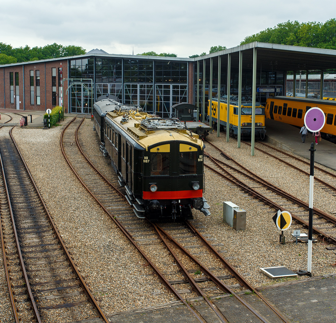 Der NS Mat ’24-Triebwagenzug (BD 9107, C 8553 und C 8104) „Blokkendozen“ (Kastendose) am 24 Juni 2025 im Het Spoorwegmuseum (bis 2005 NSM - Nederlands Spoorwegmuseum / Niederl�ndischen Eisenbahnmuseum) in Utrecht im ehemaligen Bahnhof Maliebaanstation. Ein Eisenbahnmuseum das zum Verweilen einl�dt. Der dreiteilige Zug vom Typ Mat’24 des Niederl�ndischen Eisenbahnmuseums besteht aus BD 9107, C 8553 und C 8104

Der Triebwagen BD 9107 (mBD):
Hier im Vordergrund der Triebwagen NS BD 9107 (mBD). Er wurde 1927 von Beijnes, offiziell Koninklijke Fabriek van Rijtuigen en Spoorwagens J.J. Beijnes (K�nigliche Fabrik f�r Kutschen und Eisenbahnwagen J.J. Beijnes) in Haarlem, mit einer elektrischen Ausr�stung von Metropolitan Vickers, gebaut und an die NS - Nederlandse Spoorwegen geliefert. Er hat 40 Sitzpl�tzen der zweiten Klasse sowie ein Gep�ckabteil. Seine Historischen Nummer waren ab 1927: BD 9007 / 1931: BD 9107 / 1946: CD 9107 / 1958: BD 5902 / 1966: 159016 / 1968: 9741937 / 1989 BD 9107.

Der Wagen kehrte Ende 1945 in besch�digtem Zustand aus Deutschland zur�ck. Er wurde 1947 instandgesetzt und als CD 9107 (mCD) wieder in Betrieb genommen. In den Jahren 1957/58 erfolgte der Umbau zu einem antriebslosen Wagen (BD 5902/BDz), dabei wurde die Inneneinrichtung der zweiten Klasse im „neuen Stil“ eingebaut, wobei die Anzahl von 40 Sitzpl�tzen erhalten blieb. Die elektrische Ausr�stung (einschlie�lich der Stromabnehmer) wurde entfernt, der F�hrerstand durch eine Plattform mit �bergangsfaltenbalg ersetzt und das Gep�ckabteil um einen Seitengang erweitert. Nur vier Jahre sp�ter wurde der Wagen �berfl�ssig und zur Nummer 159016, einem Schulungswagen f�r die Abteilung Gleisbau und Infrastruktur, umgebaut. Nach der Fertigstellung eines festen Schulungsraums im Jahr 1987 wurde der Wagen f�r die museale Erhaltung vorgesehen.

Die Restaurierung und der Umbau wieder zum Elektrotriebwagen BD 9107 erfolgten zwischen 1987 und 1989 im Ausbesserungswerk Amersfoort, dabei wurde das Fahrzeug weitgehend nach den Originalspezifikationen der 1930er Jahre hergerichtet, jedoch mit einer elektrischen Anlage der Firma Heemaf ausgestattet. Im Mai und Juni 2010 wurde der BD 9107 mit einer vereinfachten Version des automatischen Zugbeeinflussungssystems (ATBe) ausger�stet und im Strukton-Werk in Zutphen �berpr�ft. Die Betriebszulassung erfolgte im September, gefolgt von einer ersten Fahrt f�r die Ehrenamtlichen des Eisenbahnmuseums am 9. Oktober 2010. 

Im November 2011 wurden der BD 9107 und der Barwagen C 8553 zur �berholung der Fahrwerke in das NedTrain-Werk nach Haarlem �berf�hrt. Die eigentliche Aufarbeitung der Drehgestelle �bernahm die Firma Reuschling in Hattingen (Deutschland), dabei wurden unter anderem s�mtliche Tragfedern durch neue ersetzt. Im Dezember 2012 erhielt der gesamte Zug nach einer erfolgreichen Probefahrt im �ffentlichen Streckennetz die erneute Betriebszulassung. Der „Blokkendoos“-Zug ist aber derzeit aufgrund umfangreicher Instandhaltungsarbeiten au�er Betrieb.

Die NS-Baureihe Mat ’24 oder Materieel ’24 war eine (die erste) Serie von Elektrotriebz�gen, die von der NS - Nederlandse Spoorwegen (Niederl�ndischen Staatsbahnen, heute Niederl�ndische Eisenbahnen AG) ab 1924 in gro�er Zahl eingesetzt wurde. Diese Z�ge sind unglaublich robust und schwer. nd der Tatsache, dass die Trainer wie Bausteine zusammengestellt werden k�nnen. Im Volksmund erhielten sie wegen des eckigen �u�eren und der Tatsache, dass die Wagen wie Bausteine zusammengestellt werden konnten, den Spitznamen „Blokkendozen“ (Kastendose). Ein weiterer Spitzname war „buffermaterieel“, weil sie im Unterschied zu anderen, mehr windschl�pfig aussehenden elektrischen Triebz�gen mit �blicher Schraubenkupplung versehen waren. Wegen der Verbindung �ber Schraubenkupplung mit zwei Puffern konnten die Einheiten sehr flexibel kombiniert werden. Meist bestand ein Zug aus zwei Triebwagen und zwei oder drei antriebslosen Mittelwagen. Die lauten Ger�usche der Motoren und Antriebe erinnerten manche Fahrg�ste an Staubsauger, so dass auch dieser weitere Spitzname  Staubsauger  verwendet wurde. 

Ab Mitte der 1950er Jahre wurden die Mat ’24 zunehmend durch die neue Baureihe Mat ’54 ersetzt. Der letzte elektrische „Blokkendoos“-Zug verkehrte am 31. Mai 1959. Einige Wagen blieben als Reisezugwagen bis 1973 im Einsatz, sie wurden unter anderem in Milit�rz�gen sowie zur Kapazit�tserweiterung w�hrend der Hauptverkehrszeiten verwendet. Bei einigen Triebwagen wurden die Stromabnehmer und Motoren demontiert, sie wurden auch als einfache Beiwagen weiterverwendet. Da die Mat ’24 sehr robust waren, wurden einige Einheiten sp�ter auch als Postwagen oder f�r Dienstzwecke eingesetzt. Mehrere Wagen blieben museal erhalten.

TECHNISCHE DATEN Triebwagen BD 9107 (mBD):
Baujahr: 1927
Spurweite: 1.435 mm (Normalspur)
L�nge �ber die Puffer:	19.800 mm
Achsfolge:Bo'Bo'
Achsabstand im Drehgestell: 2.500 mm
Treibraddurchmesser: 1.000 mm (neu)
Eigengewicht: 59.000 kg
Tragf�higkeit Gep�ckraum: 2,5 t
Traktionsmotoren: 4 St�ck vom Typ Heemaf TM 8
Stundenleistung: 4 x 136 = 544 kW (Urspr�ngliche Leistung 661,5 kW)
Dauerleistung: 4 x 100 = 400 kW
�bersetzung: 27 : 56
H�chstgeschwindigkeit: 100 km/h
Kupplung: Schraubenkupplung (UIC-Kupplung)

