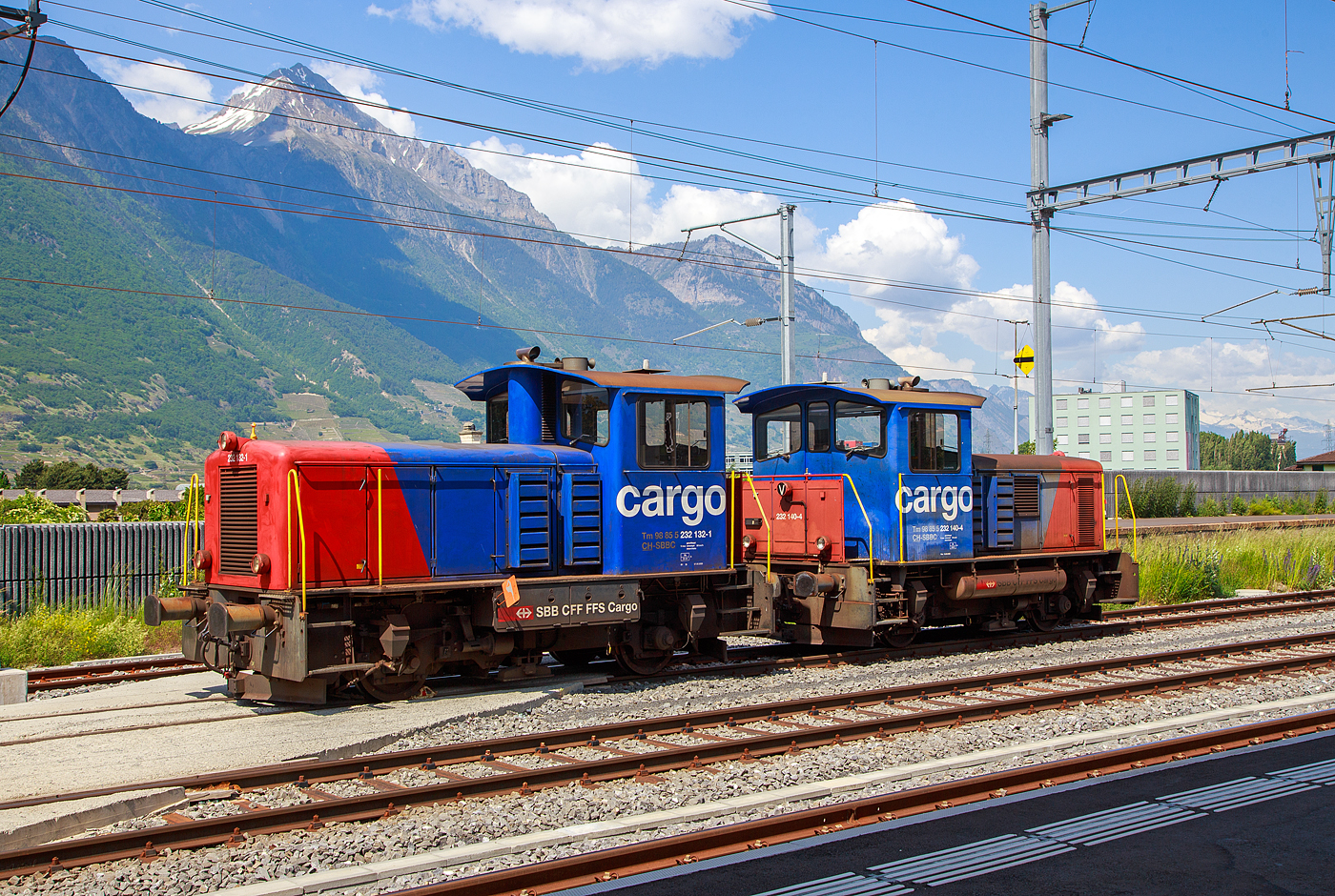 Die beiden ex SBB Tm IV Diesellok (Rangiertraktor mit Dieselantrieb), die 232 132-1 (Tm 98 85 5 232 132-1 CH-SBBC) und die 232 140-4 (Tm 98 85 5 232 140-4 CH-SBBC) sind am 26.05.2023 beim Bahnhof Martigny abgestellt.

Die Tm IV wurden 1975 von der SLM (Schweizerische Lokomotiv- und Maschinenfabrik) in Winterthur gebaut. Die 232 132-1unter der Fabriknummer 5048 als Tm 8782 und die 232 140-4 1unter der Fabriknummer 5048 als Tm 8790, jeweils f�r die SBB - Schweizerische Bundesbahnen, 2001 gingen sie an die SBB Cargo �ber. Beide wurden 2010 bzw. 2011 im Industriewerk Biel der SBB zu den heutigen Tm 232 umgebaut, es erfolgten eine Remotorisierung und der Einbau einer Funkfernsteuerung. Wobei normalerweise m�sste man sie mit der Funkfernsteuerung nun als Tmf f�hren.

Die Fahrzeuge wurden als fahrdrahtunabh�ngiges Fahrzeug f�r den Rangier- und Baudienst beschafft. Erstmals bei einer gro�en SBB-Fahrzeugserie, kam bei diesen Fahrzeugen die hydraulische Kraft�bertragung zum Einsatz. Da das Turbowendegetriebe eine Ausr�stung f�r beide Fahrtrichtungen hatte, konnte auf das st�rungsanf�llige mechanische Wendegetriebe verzichtet werden. Um die ung�nstige Wirkungsgradcharakteristik abzuschw�chen, wurde ein nur im Stillstand schaltbares Stufengetriebe eingebaut, das die Wahl zwischen einer H�chstgeschwindigkeit von 30 oder 60 km/h, oder die Wahl zwischen gr��erer Zugkraft oder gr��erer Geschwindigkeit erm�glichte.

Die Rangiertraktoren besitzen einen Au�enrahmen mit obenliegenden Blattfedern, die sich auf die Achslager abst�tzen. Die Federn besitzen keinen Lastausgleich.Die Kraft�bertragung erfolgt von der Motorwelle �ber eine Kardanwelle (Gelenkwelle) zu einem zentralen Schaltgetriebe. Dieses ist als Voith-Turbogetriebe mit nachgeschaltetem Verteilgetriebe ausgef�hrt. Von dessen Antriebswelle (beidseitig)  weiter �ber zwei Gelenkwellen auf eine Achsgetriebe, das sich auf der jeweiligen Antriebachse befindet. Die beiden Achsen sind �ber die Gelenkwellen und Antriebswellen mechanisch verbunden.

Das Voith-Turbogetriebe ist ein vollautomatisch arbeitendes hydrodynamisches Str�mungsgetriebe, das zwei Wandler besitzt. Als Betriebsfl�ssigkeit kommt Mineral�l zu Einsatz. Die beiden Wandler sind zueinander gegenl�ufig, wodurch die Fahrtrichtung ge�ndert wird. Dem Turbogetriebe ist das Verteilgetriebe nachgeschaltet. Dort kann mechanisch die �bersetzung zwischen Rangier- und Streckengang geschaltet werden. Da es sich um ein nicht synchronisiertes Schaltgetriebe handelt, darf es nur im Stillstand geschaltet werden. Es besitzt auch eine Neutralstellung, in welchen der Schalthebel bei Schleppfahrt gebracht und verriegelt wird. Das Achsgetriebe besteht aus einem Kegelradgetriebe mit einem �bersetzungsverh�ltnis 1:3.357. Auf der der Kardanwelle abgewandten Seite ist das Geh�use des Achsantriebes mit einer Drehmomentabst�tzung mit dem Fahrzeugrahmen verbunden.

Als Motor wurde ein schnelllaufender nicht aufgeladener 8-Zylinder Dieselmotor eingebaut. Die ersten Fahrzeuge waren mit einem MAN Motor des Typs R8V 16/18, mit 280 kW / 380 PS Leistung, ausger�stet, dessen Produktion sp�ter von der MTU �bernommen wurden. 

Umbau zu Tm 232
Neben der Neumotorisierung war der Einbau einer Zugsicherung der wichtigste Umbaupunkt, denn es werden doch heute vermehrt Streckenfahrten mit den Rangiertraktoren unternommen. Es kommt ein Caterpillar 6 Zylinder-Dieselmotor des Typs 13C Acert mit Ladeluftk�hler und Partikelfilter zum Einbau. Die Motorleistung betr�gt nun 310 kW, mehr l�sst das hydraulische Getriebe auch nicht zu. Daneben wurde die Spannung der Steuerung von 36 Volt auf 24 Volt abge�ndert. Neben dem Einbau einer neuen Sicherheitssteuerung und Zugsicherung werden auch alle Fahrzeuge mit einer modernen Funkfernsteuerung ausger�stet. Die Umbauten wurden im Industriewerk Biel durchgef�hrt, das heute der SBB Division Personenverkehr zugeteilt ist.

TECHNISCHE DATEN (Tm 232, ex Tm IV)
Spurweite:  1.435 mm (Normalspur)
Achsformel: B
L�nge �ber Puffer:  7.670 mm
Achsabstand:  3.570 mm
Treibraddurchmesser:   950 mm
Kleinster bef. Halbmesser:  35m
Dienstgewicht:  30 t
H�chstgeschwindigkeit: 30 (Rangiergang) / 60 km/h (Streckengang) / 90 km/h (geschleppt)
Traktionsleistung:  310 kW/421 PS (urspr. 280 kW / 380 PS)
Anfahrzugkraft: 90 kN
Dauerzugkraft:  60 kN
Motor (neu): 6 Zylinder Reihenmotor vom Typ Caterpillar 13C Acert)
Urspr�nglicher Motor:  8 Zylinder Reihenmotor vom Typ MAN Typs R8V 16/18
Leistungs�bertragung:  Hydraulisch
Tankinhalt:  850 l (Diesel)