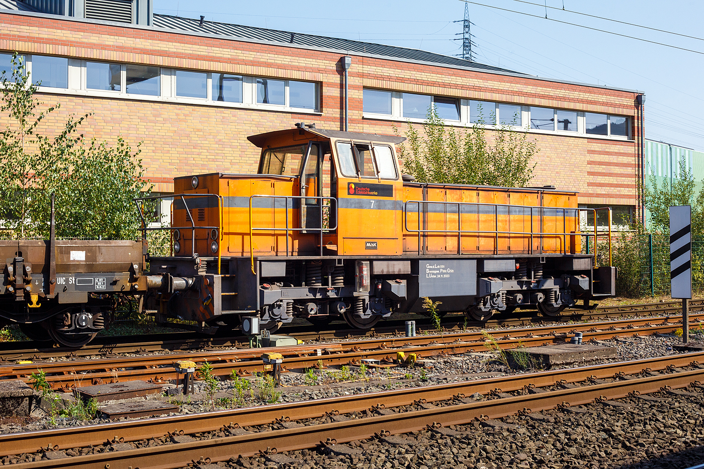 Die DEW 7 (Lok 7 der Deutschen Edelstahlwerke GmbH), zuvor Edelstahlwerke Südwestfalen, rangiert am 05 September 2023 mit vierachsigen Drehgestellflachwagen am Haken, vom Anschlussgleis zurück aufs Werksgelände in Siegen-Geisweid.

Die Lok ist eine MaK G 1203 BB und wurde1986 unter der Fabriknummer 1000808 in Kiel gebaut und an die Krupp Stahl AG, Werk Siegen-Geisweid, geliefert.

Die Lokomotive MaK G 1203 BB ist eine dieselhydraulische Lokomotive, die von der Maschinenbau Kiel (MaK) gebaut wurde, die Achsfolge ist B’B’. Sie erreicht eine maximale Geschwindigkeit von bis zu 70 km/h. Eingebaut wurden Motoren von MTU (745 kW) und für sechs Exporte nach Gabun mit amerikanischen Cummins-Motoren (761 kW). Die MaK G 1203 BB wurde zwischen 1982 und 1991 in 25 Exemplaren gebaut. Sie gingen an 14 verschiedene Eisenbahnbetreiber in Deutschland. Eine Lok ging nach Schweden und sechs nach Gabun.

Im Deutschen Fahrzeugeinstellungsregister wurde für diese Bauart die Baureihennummer 98 80 0273 vergeben.

Ab 1978 wurden im dritten Typenprogramm zwei verschiedene vierachsige Loks angeboten. Die leistungsschwächere erhielt die Bezeichnung G 1201 BB. Sie setzte sich nicht so recht durch und wurde nur einmal gebaut. Als ab 1981 die leistungsstärkeren Motoren der Reihe 396 zur Verfügung standen, änderte man die Typenbezeichnung in G 1203 BB. Äußerlich kann man diese beiden Typen an ihrem Lüftungsgitter an der Vorderseite der langen Haube erkennen.

TECHNISCHE DATEN:
Spurweite: 1.435 mm (Normalspur)
Achsfolge: B´B´
Länge über Puffer 12.500 mm
Drehzapfenabstand: 5.800 mm
Achsabstand im Drehgestell: 2.400 mm
Treibraddurchmesser: 1.000 mm (neu)
größte Breite: 3.100 mm
größte Höhe über Schienenoberkante: 4.220 mm
kleinster befahrbarer Gleisbogen: 60 m
Dienstgewicht: 88 t
Kraftstoffvorrat: 2.500 l
Motor : MTU, Typ 8V 396 TC 13, ein V8-Zylinder-Dieselmotor mit Abgasturbolader und Ladeluftkühlung,
Leistung: 745 kW (1.012 PS)
Drehzahl: 1.800 U/min
Getriebe: Voith, Typ L4r4zU2

Die Deutschen Edelstahlwerke sind die deutsche Produktionsgesellschaft der Swiss Steel Group mit Hauptsitz in Luzern, Schweiz, einem der weltweit führenden Produzenten von Spezialstahl-Langprodukten. Dank des ausschließlichen Einsatzes von Stahlschrott in Elektrolichtbogenöfen zählt die Gruppe zu den europaweit relevantesten Unternehmen in der Kreislaufwirtschaft und ist im Bereich nachhaltig produziertem Stahl – Green Steel – unter den Marktführern. Swiss Steel Group verfügt über eigene Produktions- und Distributionsgesellschaften in über 30 Ländern. 

Die Eckdaten der Geisweider Eisenwerke (heute Teil der Deutschen Edelstahlwerke) :
1845: Gründung des Stahlwerks durch J.H. Dresler als Eisen- und Walzwerk
1872: Errichtung von Hochofen 1
1879: Umbennung in  Geisweider Eisenwerke AG 
1889: Inbetriebnahme des ersten Siemens-Martin-Stahlwerks
1902: Errichtung von Hochofen 2 (mit der noch existierenden Gießhalle)
Ende 1. Weltkrieg: Stillegung beider Öfen (mehrjährige Pause wegen Weltwirtschaftskriese und Quotenvorgaben für Stahlunternehmen)
1920: Aufkauf durch Thyssen und Klöckner, anschließend Teil der Vereinigten Stahlwerke AG, dem zweitgrößten Stahlproduzenten weltweit
1938: Hochofen 2 wird renoviert und modernisiert und geht wieder in Betrieb, Stillstand am Ende des 2. Weltkriegs
1948: der verbliebene Hochofen geht wieder in Betrieb
1951: auf Annordnung der Alliierten Hohen Kommission erfolgte die Zerschlagung der VESTAG und Geisweid Teil der Stahlwerke Südwestfalen AG im Verbund mit dem Stahlwerk Hagen (vorm. Eicken), den Stahlwerken Brünninghaus und der FriedrichThomée AG aus Werdohl
1972: 100 Jahre Hochofenbetrieb in Geisweid, 70 Jahre Hochofen 2 
1974: Übernahme durch Krupp (Krupp Edelstahlprofile)
1978: Installierung eines 140 t Elektrolichtbogenofen (der größte in Deutschland)
1979/1981: Stillegung/Abbruch des Hochofens 2, es war der letzte Siegerländer Hochofen
2004: das Geisweider Werk geht an die Deutschen Edelstahlwerke in der Schmolz&Bickenbach-Gruppe zur Swiss Steel Group gehörend, wieder unter dem Namen Deutsche Edelstahlwerke Specialty Steel. 

Das Stahlwerk in Siegen-Geisweid betreibt einen 140 t Elektrolichtbogenofen und eine 6-adrige Stranggießanlage, um Edelstahl-Knüppel für das eigene Stabstahlwalzwerk und die Drahtstraße in Hagen herzustellen. Der Edelstahl wird aus Schrott hergestellt. Von einstmals rund  7.000 Arbeitsplätzen sind heute noch rund 1.100 verblieben.
