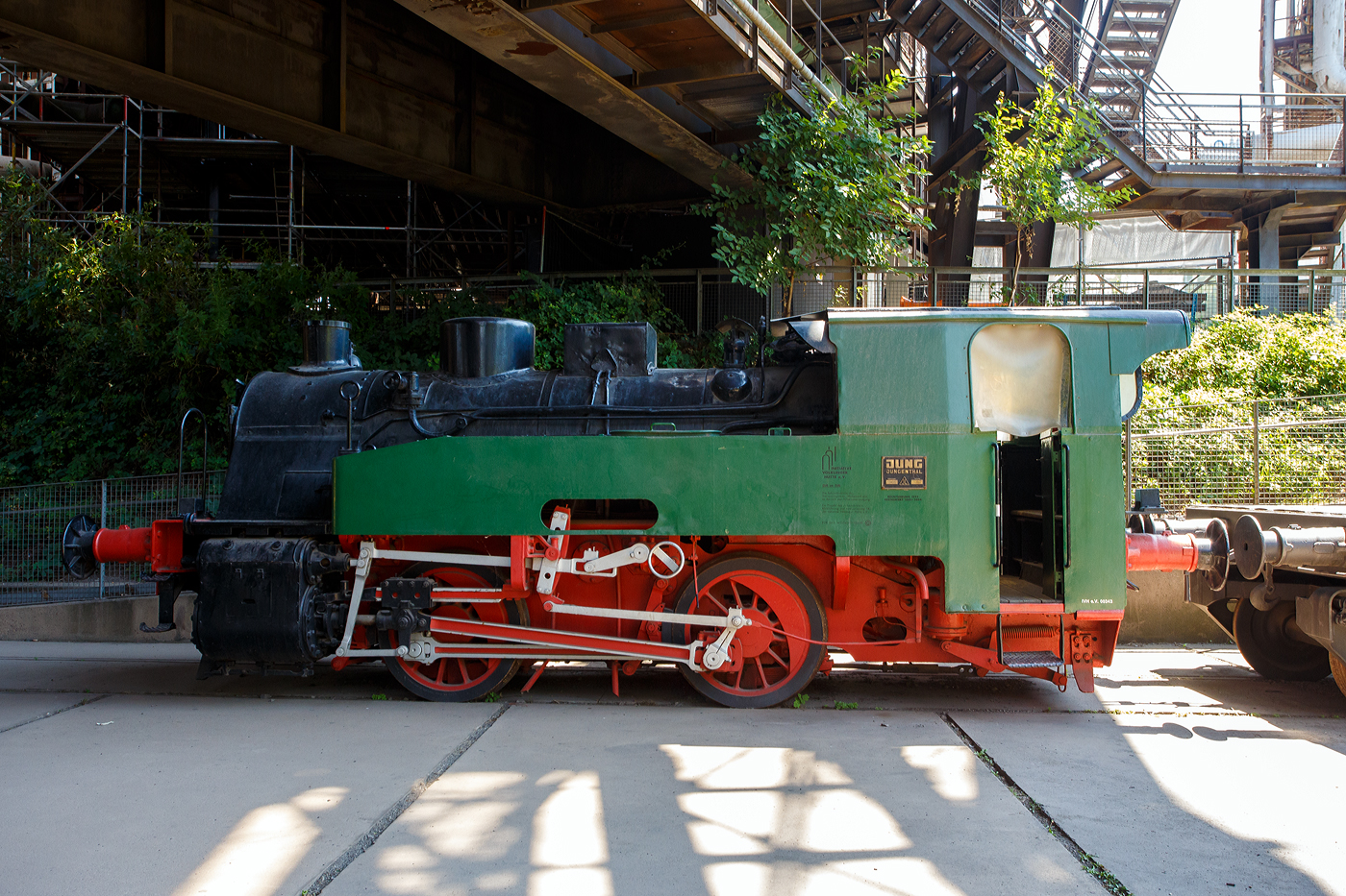 Die Normalspur-Dampflokomotive 46 der V�lklinger H�tte (ehem. R�chling'schen Eisen- und Stahlwerke GmbH in V�lklingen/Saar, ab 1982 ARBED-Saarstahl GmbH, ab 1986 Saarstahl V�lklingen GmbH), eine Jung Hochofenlok niedriger Bauart, abgestellt mit einem offenen Roheisenpfannenwagen der Burbacher H�tte, hier am 01 Juli 2025 am Gel�nde V�lklinger H�tte als Denkmal. Heutiger Eigent�mer ist die Initiative V�lklinger H�tte e.V..

1964 wurde die Lok Nr. 46 mit der Fabriknummer 13835 im Auftrag der R�chling'schen Eisen- und Stahlwerke als letzte rostgefeuerte Dampflok der Firma Arnold Jung, Lokomotivfabrik GmbH, in Jungenthal bei Kirchen an der Sieg gebaut und am 09.12.1964 geliefert. Einsatzgebiet war der Roheisentransport von den sechs Hoch�fen zum Thomasstahlwerk, wobei die besonders beengten Verh�ltnisse auf der V�lklinger H�tte eine besondere Bauform erforderten. Die Verbindung zwischen Eisenwerk und Stahlwerk war nur durch einen Tunnel m�glich, da die Deutsche Bahn durch ihre Gleisanlagen die H�tte in zwei Bereiche teilt. Dieser Tunnel, der Roheisenkanal, ist so niedrig, dass die zweiachsige Lok nur eine H�he von 2.640 mm �ber der Schienenoberkante und eine Breite von 2.450 mm aufweist. Die Loks 44 (11945/Baujahr 1953) und 45 (13240/Baujahr 1959), die nach vorliegenden Unterlagen technisch baugleich sind, wurden schon vorher (von der Arnold Jung GmbH an die V�lklinger H�tte geliefert. 

Um speziell die F�hrerhausr�ckwand, die Lampen, aber auch die Kupplungselemente zwischen Lok und dem ersten Wagen vor fl�ssigen gl�henden Spritzern von herabfallenden Roheisen zu sch�tzen, erhielten die Lokomotiven ein nach hinten verl�ngertes Dach. Die Puffer wurden im Stahlwerk den dort vorhanden Roheisenpfannenwagen angepasst. Alte Jung Mitarbeiter berichten, an der Lok h�tte man kaum was verdient, weil l�nst nicht mehr Dampflokteile ab Lager vorhanden waren umd z.T. m�hsam und teuer beschafft werden musste.

Im Zuge der Modernisierung und Rationalisierung wurden die Roheisendampfloks 1981 nicht mehr ben�tigt. W�hrend die Lokomotiven 44 und 45 an ein Museum im Ruhrgebiet verkauft wurden, gelangte die Lokomotive 46 als technisches Denkmal 1982 zur Universit�t Kaiserslautern. Diese schenkte die Lokomotive 46 der Initiative V�lklinger H�tte, die sie mit Hilfe von Sponsoren am 9. Februar 1999 nach V�lklingen zur�ckholte.

TECHNISCHE DATEN:
Baujahre: 1953,1959 und 1964
Gebaute Anzahl: 3
Spurweite: 	1.435 mm (Normalspur)
Achsformel: B
L�nge: 7.600 mm
Achsabstand: 2.000 mm
Triebraddurchmesser: 1.000 mm (neu)
Breite: 2.450 mm
H�he: 2.650 mm
Dienstgewicht: 26 t
H�chstgeschwindigkeit: k. A. 
Leistung: 250 PS
Zugkraft: 7,5 t
Zylinder: 2 St�ck, � 380 mm x 500 mm Hub
Dampfdruck: 14 bar
Heizfl�che: 58,92 m�
Rostfl�che: 1,17 m�
Anzahl Siederohre: 178 St�ck
Durchmesser Siederohre: 33/38 mm
L�nge Siederohre: 2.540 mm
Wasservorrat: 2.500 l (2,5 m�)
Kohlenvorrat: 500 kg