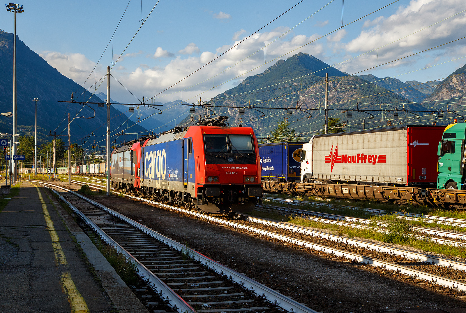 Die SBB Cargo Re 484 017-9 / E 484.017 SR (91 85 4484 017-9 CH-SBBC) und die an die SBB Cargo International AG vermietete Vectron 193 478-5  Gottardo  (91 80 6193 478-5 D-SIEAG) rollen, am 02 August 2019 im Rbf Domodossola, nun mit abgesenkten Stromabnehmern, in den FS Gleichstrombereich, zur�ck und werden sp�ter in der Abstellgruppe geparkt. 

W�hrend hinten nun zwei BLS Re 485 (TRAXX F140 AC1) den RAlpin-RoLa-Zug aus Novara nach Freiburg im Breisgau �bernehmen. Eigentlich k�nnten auch die SBB-Loks den Zug durch den Simplon und die Schweiz ziehen, die Zulassungen haben sie und die 193er sogar f�r Deutschland, �bernehmen die zwei BLS Re 485 (TRAXX F140 AC1) den Zug. Man setzt wohl auf Arbeitsteilung. Aber lange kann man dieses Schauspiel in Domodossola nicht mehr sehen, Ende 2025 wird die Rollende Autobahn eingestellt.

Diese Rola-Z�ge sind (bald waren) f�r den Transport von Last- und Sattelz�gen im begleiteten Verkehr, einer Form des kombinierten Ladungsverkehrs, vorgesehen und kommt in alpenquerenden Shuttlerelationen zum Einsatz. Begleiteten Verkehr hei�t, dass der ganze Sattelzug oder Anh�ngerz�ge mit max. 44 t und einer max. L�nge von 18,60 Meter (in Ausnahmef�llen 19,0 m), mit der Zugmaschine, auf den Zug verladen werden. Dabei fahren die Lastkraftwagen in speziellen Terminals direkt von der Stra�e �ber eine Rampe auf den Wagen, um vom Ausgangsort zum Ziel transportiert zu werden, auch der LKW-Fahrer f�hrt mit. Die Fahrer ziehen sich in einen speziellen Begleitwagen mit Liegeabteilen (Bmc) zur�ck. 

Am Ziel-Terminal angekommen �bernimmt jeder Fahrer wieder seinen LKW und f�hrt diesen wieder �ber eine Rampe vom Zug auf die Stra�e und liefert die Ware sicher beim Kunden ab. Ein gro�er Vorteil ist auch bei den Fahrern die Einhaltung der „Lenk und Ruhezeiten“. Bei kurzen Shuttlezugverbindungen als Lenkpause gewertet und auf den Mittel- und Langstrecken einen Teil oder sogar als gesamte Ruhezeit gewertet. Dabei trotzdem n�her und ausgeschlafen an Ihr Ziel. Zudem gibt es f�r die Fahrer ein On-Board Service mit Speisen und Getr�nken. 

Weitere Vorteile sind:
•	die Reduzierung des Treibstoffverbrauches und Betriebskilometer
•	die Einsparung der diversen Mauten und Sondermauten
•	die Steigerung des Umsatzes und der Produktivit�t
•	den Einsatz des Lkw trotz Wochenend-, Feiertags-, Nacht- und sonstiger Fahrverbote
•	ersparen von Stauwartezeiten, �berholverbote und andere Behinderungen
•	die Verbesserung der �KO-Bilanz des Unternehmens (CO2-Ersparnis)
•	hohe Sicherheitsstandards der Z�ge
•	Einhaltung von Lenk und Ruhezeiten der Fahrer (allein) auch bei gro�en Entfernungen

Diese Form des kombinierten Verkehrs von Schiene und Stra�e – Rollende Landstra�e (ROLA) – gewann, nicht zuletzt aufgrund von gesteigertem Verkehrsaufkommen und Umweltbewusstsein, immer mehr an Bedeutung. In �sterreich bleibt es wohl auch so, aber hier Italien – Schweiz – Deutschland ist leider Ende 2025 Schluss damit, sehr schade. Obwohl die RAlpin noch Ende 2023 vermeldet hat: knapp 12% mehr Lastwagen per Bahn
umweltfreundlich durch die Schweizer Alpen transportiert als im Vorjahr. 

Die Medienmitteilung RAlpin AG vom 05. Mai 2025 (komplett zitiert):
Die ‚Rollende Autobahn’ wird Ende 2025 eingestellt.

Wegen unerwartet vielen Einschr�nkungen auf dem Schienennetz kann das
Unternehmen RAlpin die ‚Rollende Autobahn‘ (Rola), d. h. den Bahnverlad von ganzen Lastwagen f�r die Fahrt durch die Alpen, auch mit den Finanzhilfen des Bundes nicht mehr wirtschaftlich betreiben, obwohl die Nachfrage von Seiten der Kunden weiterhin gut ist. In Absprache mit dem Bund wird die Rola deshalb fr�her eingestellt als geplant, n�mlich per Ende 2025 statt wie bisher vorgesehen Ende 2028.

Die RAlpin AG steht vor erheblichen wirtschaftlichen Herausforderungen. Trotz der laufenden finanziellen Abgeltungen des Bundes, einer vorhandenen Nachfrage und guter Auslastung von 80 Prozent ist der Betrieb der ‚Rollenden Autobahn’ (Rola) nicht mehr wirtschaftlich m�glich. Bereits 2024 fielen rund 10 Prozent der Z�ge aus. Der Grund waren geplante sowie kurzfristig angeordnete Baustellen und weitere unvorhersehbare Ereignisse. Dies f�hrte 2024 zu einem negativen Ergebnis von rund -2.2 Millionen Franken. Im 1. Quartal 2025 konnten wegen Bauarbeiten im Vergleich zum Vorjahr rund 20 Prozent weniger Z�ge gefahren werden; statt 1018 Z�ge im 1. Quartal sind dieses Jahr in der gleichen Zeitperiode nur deren 794 gefahren.

Das eidgen�ssische Parlament hatte vor zwei Jahren beschlossen, die finanzielle F�rderung der Rola durch den Bund ein letztes Mal zu verl�ngern und den Betrieb per Ende 2028 einzustellen. Die RAlpin kommt nun zum Schluss, dass die Rola unter den ge�nderten Voraussetzungen nicht mehr wirtschaftlich betrieben werden kann, zumal eine Normalisierung der Situation nicht in Sicht ist. In Absprache mit dem Bund hat sie deshalb beschlossen, den Betrieb bereits auf den im Dezember 2025 einzustellen. Die drei Aktion�re der RAlpin (BLS, Hupac und SBB) sind bereit, bis dahin die Finanzierung der Rola sicherzustellen, um eine geordnete Betriebseinstellung zu erm�glichen und die gesch�ftlichen Verpflichtungen zu erf�llen. Der Bund erh�ht seinerseits bis zur Einstellung der Rola die durchschnittliche Abgeltung je verlagerten Lastwagen.

Bei RAlpin sind 16 Personen angestellt, die RAlpin sucht nach L�sungen f�r die betroffenen Mitarbeitenden. Da bereits seit l�ngerem klar ist, dass der Betrieb bis maximal Ende 2028 weitergef�hrt wird, ist RAlpin im steten Austausch �ber den individuellen k�nftigen Berufsweg von allen Mitarbeitenden.

Die Herausforderungen im alpenquerenden kombinierten Verkehr sind gro�:
Die RAlpin hat in den letzten Jahren die notwendigen Voraussetzungen geschaffen, trotz Kostensteigerungen f�r die Nutzung der Bahninfrastruktur, Energie, Terminals und Traktion mit einem konstanten Zahlungsrahmen j�hrlich bis zu 80.000 Lastwagen im begleiteten kombinierten Verkehr durch die Alpen auf die Schiene zu verlagern. Dies entspricht 7 Prozent des kombinierten Verkehrs durch die Schweizer Alpen. Die RAlpin wird gemeinsam mit den Kunden M�glichkeiten suchen, um ihre Mengen weiterhin auf der Schiene zu transportieren. Es ist aber davon auszugehen, dass ein Teil, der auf der Rola transportierten G�ter zuerst auf die Stra�e zur�ck verlagert wird, bis die Umr�stung auf kranbare Sattelauflieger erfolgt ist. Wobei es gibt Systeme bei denen die Sattelauflieger nicht kranbar sein m�ssen.

Die Situation der Rola ist symptomatisch f�r den ganzen alpenquerenden kombinierten Verkehr auf der Schiene. Grund f�r die zunehmenden Zugausf�lle auf der Rola ist insbesondere die anhaltend hohe St�rungsanf�lligkeit der Schieneninfrastruktur in Deutschland. Die RAlpin erwartet, dass sich die Bedingungen f�r die Akteure im kombinierten Verkehr in den kommenden Jahren verbessern werden und so neue tragf�hige L�sungen m�glich sind, um das Verlagerungsziel von der Stra�e auf die Schiene zu erreichen. Dazu wird es weiterhin flankierende Ma�nahmen durch die Politik brauchen. Es muss das Ziel sein, die Anstrengungen der letzten 25 Jahre f�r eine erfolgreiche Verlagerung des alpenquerenden Verkehrs durch die Schweiz auf die Schiene langfristig zu sichern.

So soll langfristig der unbegleitete kombinierter Verkehr (ohne LKW-Zugmaschine) die RoLa abl�sen: 
Die ‚Rollende Landstrasse’ (Rola) war seit den sp�ten 1960er-Jahren das erste Angebot, um den G�terschwerverkehr durch die Alpen von der Stra�e auf die Schiene zu verlagern. Dabei werden ganze Lastwagen oder Sattelschlepper auf die Schiene verladen und die LKW-Chauffeure reisen in einem Begleitwagen im Zug mit. Daher ist die Rola auch als „begleiteter kombinierter Verkehr“ bekannt.

Im Zuge des Ausbaus der Alpentransitstrecken wurde die Rola 2001 mit der neu gegr�ndeten RAlpin AG, einem Gemeinschaftsunternehmen von SBB, BLS und Hupac, modernisiert. Die Betreiberin transportiert Lastwagen auf der Strecke zwischen Freiburg im Breisgau (D) und Novara in Italien. Das Angebot war von Beginn weg als �bergangsl�sung konzipiert, bis die Fertigstellung der NEAT einen effizienten Bahntransport von Sattelaufliegern (Sattelschlepper ohne Zugfahrzeug und Chauffeur) und Containern �ber lange Distanzen erlaubt. Das eidgen�ssische Parlament hatte vor zwei Jahren beschlossen, die finanzielle F�rderung der RoLa durch den Bund ein letztes Mal zu verl�ngern und den Betrieb per Ende 2028 einzustellen.

Parallel dazu entwickelte sich der „unbegleitete Kombinierte Verkehr“ dank fortschrittlichen Umschlagtechniken wie kranbaren Sattelauflegern und Containern weiter. Wobei bei es gibt heute schon Systeme bei denen nicht kranmanipulierbare Sattelauflegern auch im KLV gefahren werden k�nnen. Erw�hnenswert sind da u.a. das System „NiKRASA“ der TX Logistik AG, das weltweit patentierte System Trailer-Wagen der Helrom GmbH, sowie das horizontale Verladesystem der CargoBeamer AG. Mit der Fertigstellung der NEAT geh�rt die Zukunft auf dem Nord-S�d-Korridor dieser Sparte des kombinierten Verkehrs.

RAlpin AG auf einen Blick
Die RAlpin AG mit Sitz in Olten betreibt die Rollende Autobahn (Rola) zwischen Freiburg i. Br. und Novara. Ihr Angebot umfasst j�hrlich 100.000 Verladegelegenheiten f�r Lastwagen von der Stra�e auf die Schiene. Im 2024 wurde das Angebot von rund 72.000 Lastwagen genutzt. Die Lastwagenfahrer/-innen verladen ihre Fahrzeuge in den Terminals auf Niederflurtragwagen und reisen sicher in einem Begleitwagen mit. Mit diesem Angebot leistet RAlpin einen Beitrag zur Verkehrsverlagerung von der Stra�e auf die Schiene und zum Schutz der sensiblen Alpenregion. (Zusatz von mir: Nur leider spielt Deutschland nicht so mit).

Die Aktion�re der RAlpin sind BLS AG, Hupac SA und SBB AG.

Quellen: RAlpin, Railcargo
Stand: September 2025
