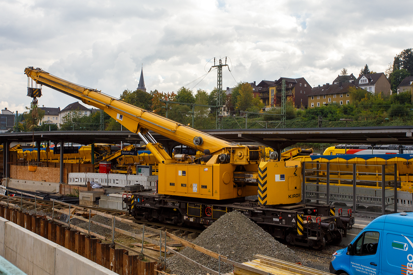 Nun im Einsatz der 125 t KIROW Gleisbauschienenkran KIROW Multi Tasker KRC 810 T (mit Schwerlastausrüstung), Schweres Nebenfahrzeug Nr. D-HGUI 99 80 9419 024-1, ex 97 82 53 513 19-3, der HERING Bahnbau GmbH (Burbach) ist am 29 September 2025 im Betzdorf/Sieg. 

Hier im Bereich unter dem P+R Parkhaus bis zum Fluss Sieg werden Horizontalbohrungen gemacht, vermutlich für erforderliche Entwässerungs-Leitungen. So mussten Bohrrohre in die Baugrube gehoben werden und wie hier der Abraum aus der Baugrube gehoben werden. Eigentlich ist ein 125 t Kran damit unterfordert (überdimensioniert), aber man muss bedenken es wurde eine weite Auskragung (Ausladung vor Puffer) und das freistehend (Nichtabgestützt) benötigt. 

Der hier mit der Ausladung vor Puffer von über 15 m könnte der Kran freistehend 30 t heben, aber darf dann nicht schwenken. Wenn geschenkt werden muss liegt diese dann nur bei 1,5 t. Entscheidend für die Ausladung des Auslegers ist aber auch die hintere Ausladung des Gegengewichts. 

Der Kran wurde 2008 von KIROW in Leipzig unter der Fabriknummer 149000 gebaut und an die HERING Bahnbau GmbH (Burbach) geliefert.

Der Gleisbauschienenkran KRC 810 T ist ein Schienendrehkran des Herstellers Kirow Ardelt (bis 2008 Kirow) in Leipzig. Der Typ 810 wird für den Aus-und Einbau von schweren Betonweichen, Gleisjochen und Hilfsbrücken sowie für die Montage von Ingenieurbauwerken eingesetzt. Bei Havarien auf Bahnstrecken leistet er schnelle Hilfe zum Bergen schwerer Lasten. Das maximale Lastmoment des Krans liegt zwischen 500 und 900 tm. Die Krane werden auch als Eisenbahndrehkran „EDK“ bezeichnet.

Die maximale Tragfähigkeit des bis zu 128 Tonnen schweren Krans beträgt abgestützt bis zu 125 Tonnen und freistehend 90 Tonnen. Bei maximaler Ausladung kann der Kran noch 28,5 Tonnen heben. Die größte mögliche Hakenhöhe liegt bei 18 m und die maximale Ausladung vor Puffer bei 15 m. Der Teleskoparm erlaubt Arbeiten im Radius zwischen 6,5 und 24,5 Metern. Im Gegensatz zu früheren Kranen ist auch ein Einsatz in der Nullposition ohne Abbau der Fahrleitung möglich. Die Gegenlast (Gegengewicht) ist zum Ausgleich unterschiedlicher Lasten ausfahrbar. Die Gegenlast kann in Transportstellung nicht (wie z.B. bei dem KRC 910) auf einem Gegenlastwagen abgestützt werden. Dadurch hat KRC 810 auch eine höhere Streckenlast (als z.B. der KRC 910). Für Hebearbeiten instehender Stellung verfügt der Kran über hydraulisch-ausfahrbare Stützen. Die Höchstgeschwindigkeit 19 km/h mit dem Eigenantrieb und 120 km/h im Zugverband geschleppt.  Zu dem Kran gehören zwei Kranschutzwagen. Die zulässige Anhängelast beträgt 80 t.

TECHNISCHE DATEN:
Spurweite: 1.435 mm (Normalspur)
Anzahl der Achsen: 8
Achsfolge: [(1A)(1A)][(A1)(A1)]
Länge über Puffer: 13.000 mm (ohne Ausleger)
Drehzapfenabstand: 8.000 mm
Drehzapfenabstand im Drehgestell 1 und 2: 2.300 mm
Achsabstand in den Einzeldrehgestellen: 1.100 mm
Ergebene Achsabstände in m: 1,1 / 1,2 / 1,1/ 4,6 / 1,1 / 1,2 / 1,1
Treib- und Laufraddurchmesser: 730 mm (neu) / 680 mm (abgenutzt)
Höhe in Transportstellung: 4.250 mm
Breite in Transportstellung: 3.100 mm
Eigengewicht: 128 t
Dieselmotor: Wassergekühlter 6-Zylinder Reihenmotor mit Abgasturboaufladung und Ladeluftkühlung vom Typ Cummins M 11 – C 315
Motorleistung: 235 kW bei 1.800 U/min
Max. Drehmoment: 1.458 Nm bei 1.300 U/min
Not-Dieselmotor: Luftgekühlter 2-Zylinder Viertakt-Dieselmotor mit Direkt-Einspritzung vom Typ Hatz 2 G 40 mit einer Leistung11,5 kW bei 2.300 U/min.
Fahrgeschwindigkeit im Zugverband: 120 km/h (geschleppt)
Fahrgeschwindigkeit mit Eigenantrieb: 20 km/h
zul. Anhängelast: 80 t
Hakenhöhe (max.): 18 m
Ausladung (max. vor Puffer): 16 m
Zugelassen als Schwerwagen für Streckenklasse: CS 05
Maximale Radsatzlast: 16 t
Streckenlast (Meterlast):  9,9 t/m
kleinster befahrbarer Radius: 00 m
Bremse: KE-GP-D- m.Z. (Scheibenbremsen auf allen Rädern)
Sekundär Scheibenbremsen auf allen Rädern
Handbremse: hydro-mechanisch auf alle Radsätze
Kraftstoffvorrat: 990 l

Tragfähigkeiten:
bei 0,00 m Ausladung vor Puffer, mit Schwerlastausrüstung: 125 t
bei 2,80 m Ausladung vor Puffer, ± 30°, abgestützt: 90 t
bei 2,80 m Ausladung vor Puffer, ± 20°, freistehend: 46 t